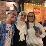 Three female students lean together and smile for the camera inside a local pizza restaurant.