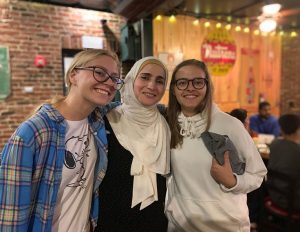 Three female students lean together and smile for the camera inside a local pizza restaurant.