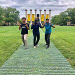 Three students pose with their hands like tiger claws in front MU's six columns on a grassy quad.