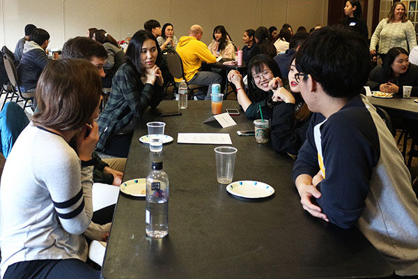 Asian and domestic students sit along both sides of a long table in a crowded room.
