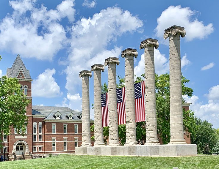 Three american flags hang between the stone columns on the grassy quad.