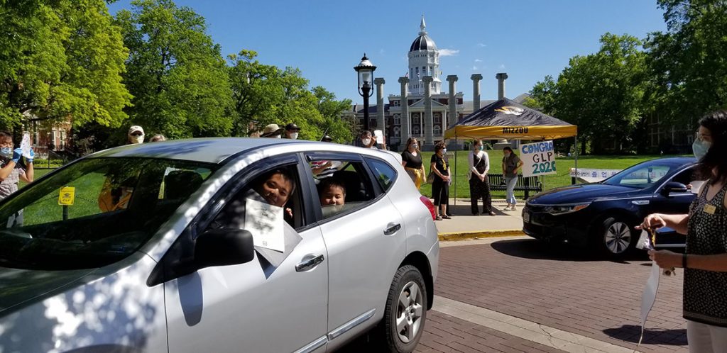 A scholar smiles and holds his certificate out his car window as he drives through the graduation celebration.