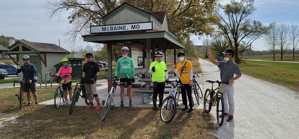 Scholars and staff pose with their bikes alongside a gravel trail; a covered shelter with a sign that reads 'McBaine, MO' is in the background
