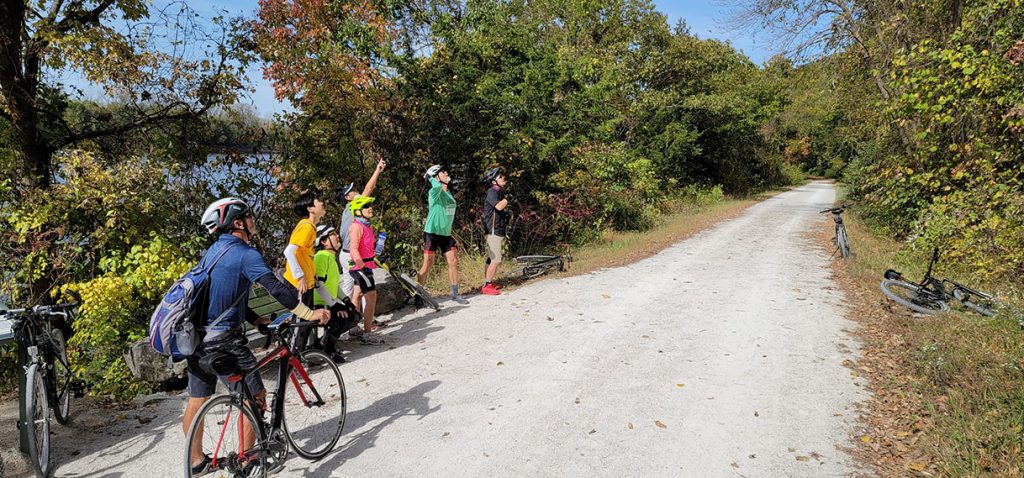 Scholars and staff stand and sit with bikes alongside the gravel trail looking up at something out of frame