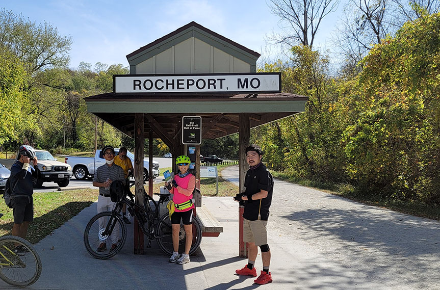 Scholars and staff stand with bikes alongside a gravel trail and under a covered shelter with a sign that reads 'Rocheport, MO'