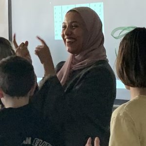 Photo of Sohaila Bakr, smiling and gesturing with her hands as she gives a presentation to elementary school students, who are facing towards her and away from the camera.