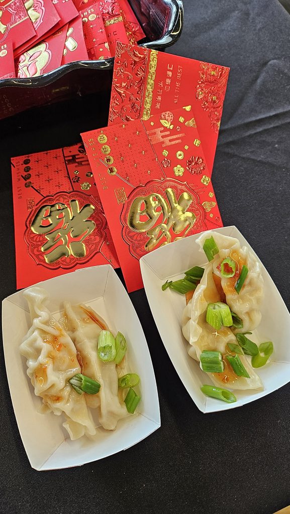 Dumplings in paper trays and red envelops sit on a black cloth covered table.