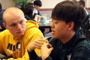 Two male students sit together at a table and talk.