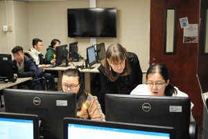 An instructor stands, helping two students seated at computers in a lab.