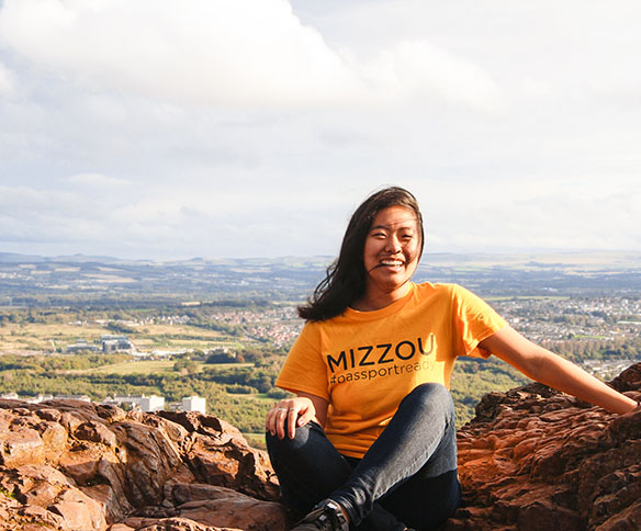 A student wearing a gold Mizzou shirt sits on the rocky top of Arthur's Seat, overlooking Edinburgh.